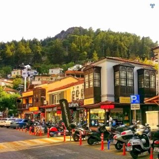 A historic high street lined with Greek Ottoman buildings. A steep wooded rocky crag backs the street.