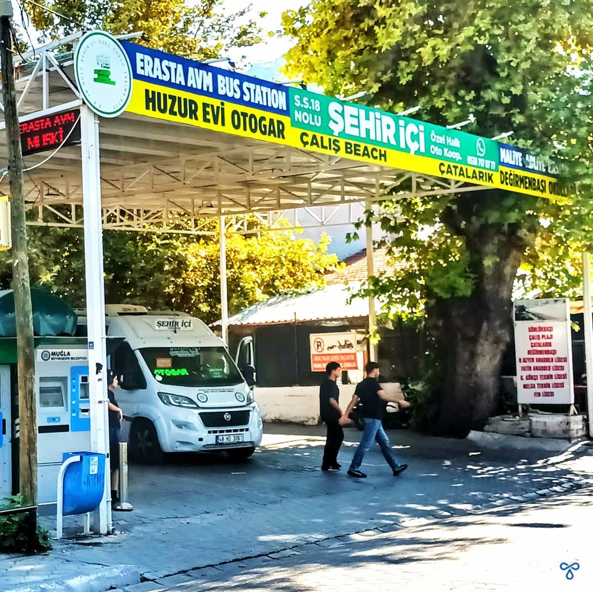 Bus station in Fethiye with white minibus, waiting for passengers. Destinations are written on a banner along the roof of the stand.