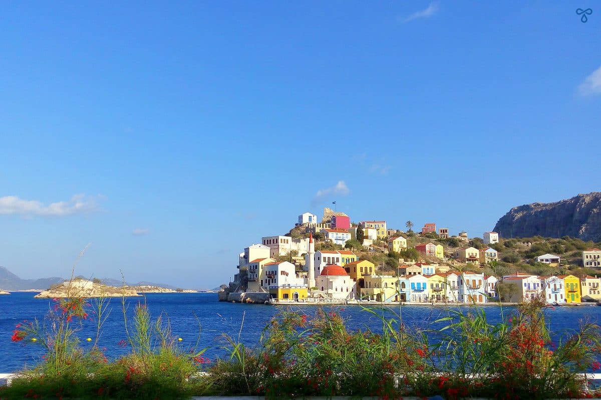 A view across the harbour of Kastellorizo. Colourful buildings, including a mosque and a castle, climb up the hillside.