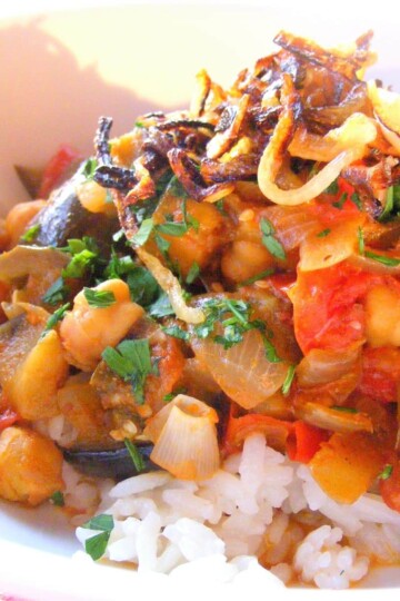 Chickpeas, aubergine chunks and crispy fried onion top a bed of rice in a white bowl.