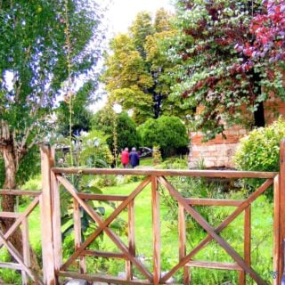 Two people walking through mature gardens. A wooden fence is in the foreground.