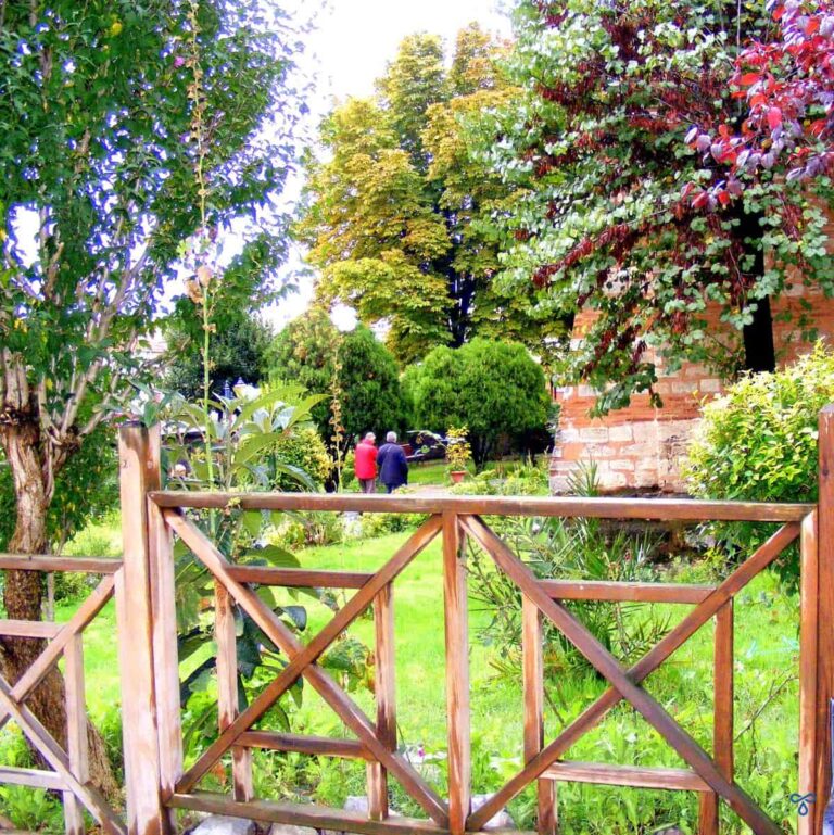 Two people walking through mature gardens. A wooden fence is in the foreground.