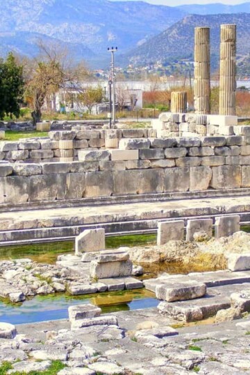 Temple Of Leto At Letoon, Turkey. There are two columns and a long base to the rear. The temple is surrounded by water.