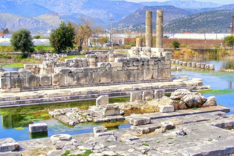 Temple Of Leto At Letoon, Turkey. There are two columns and a long base to the rear. The temple is surrounded by water.