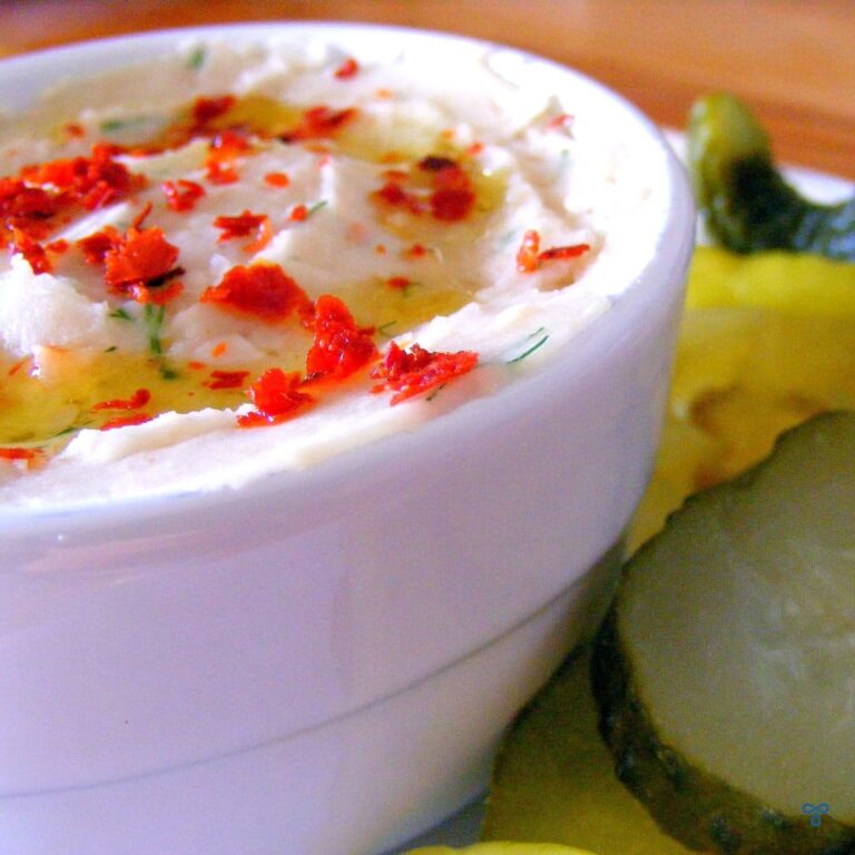 A white ramekin dish filled with a white bean dip and topped with olive oil and chill flakes. Pickled vegetables are on the side.