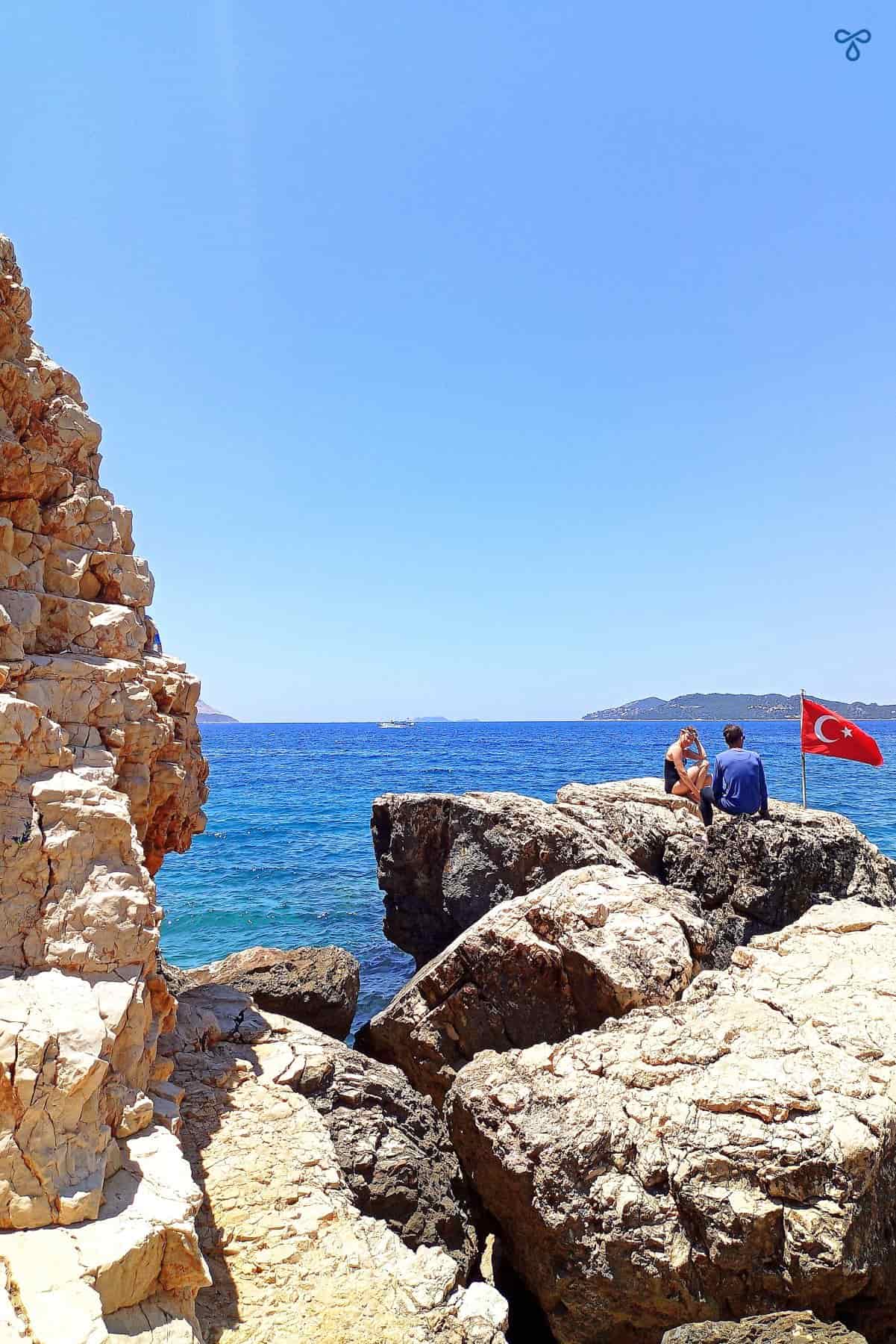 A couple sitting on rocks overlooking the sea. A Turkish flag flies next to them.