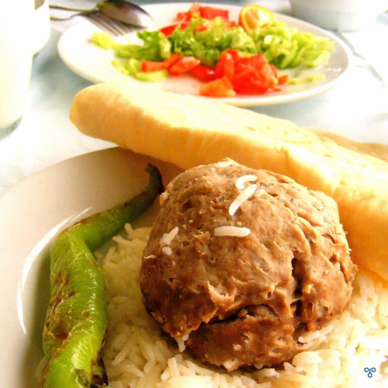A large meatball sits on a bed of rice. A long green pepper and a roll of lavash bread are either side of the meatball. A small side salad is in the background.