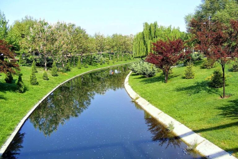 A still river curves through a manicured park with lawn and shrubs either side.