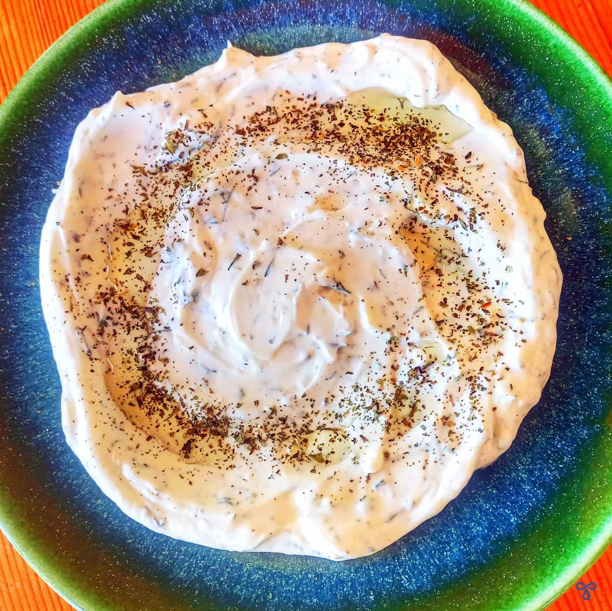 A blue & green plate with a portion of haydari yoghurt dip. The dip is topped with olive oil and dried mint.