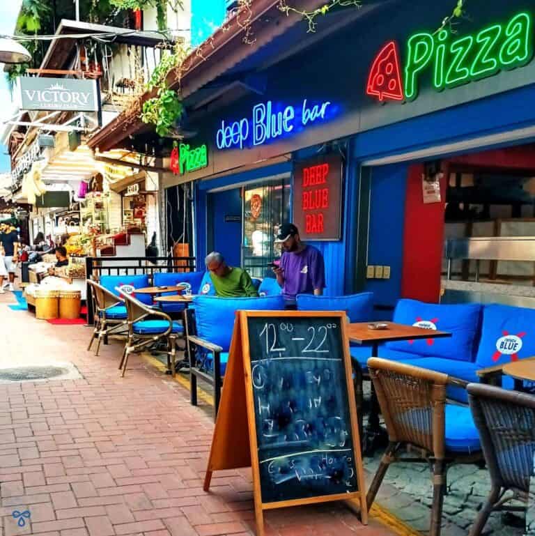 A street view outside Deep Blue Bar in Fethiye. A board advertises drink prices and a neon sign advertises pizza.