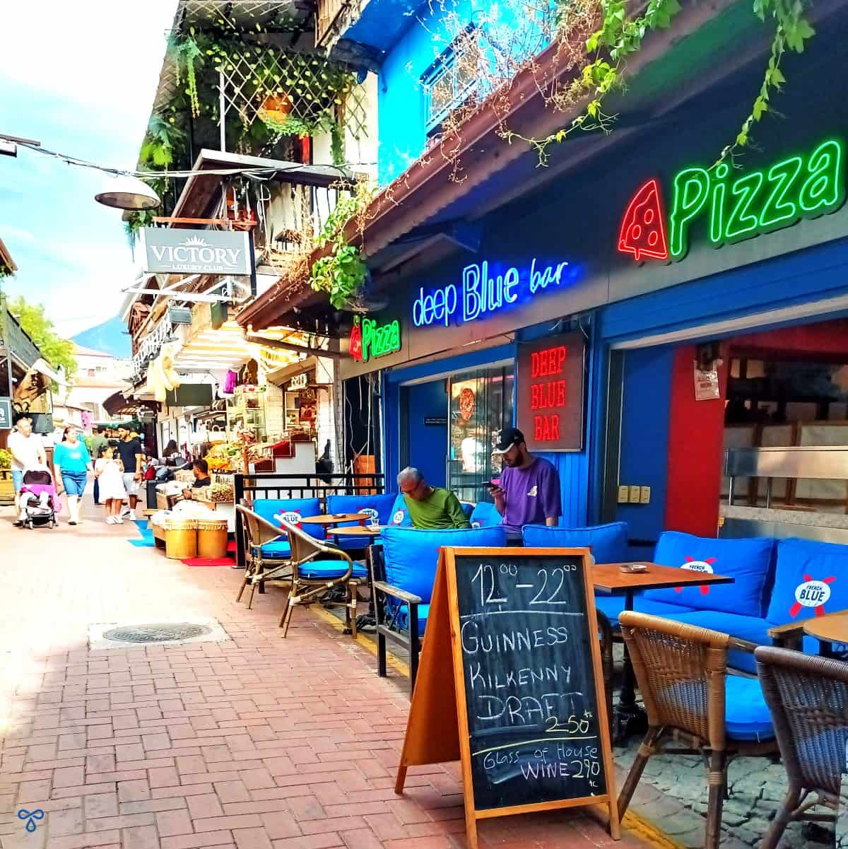 A street view outside Deep Blue Bar in Fethiye. A board advertises drink prices and a neon sign advertises pizza.