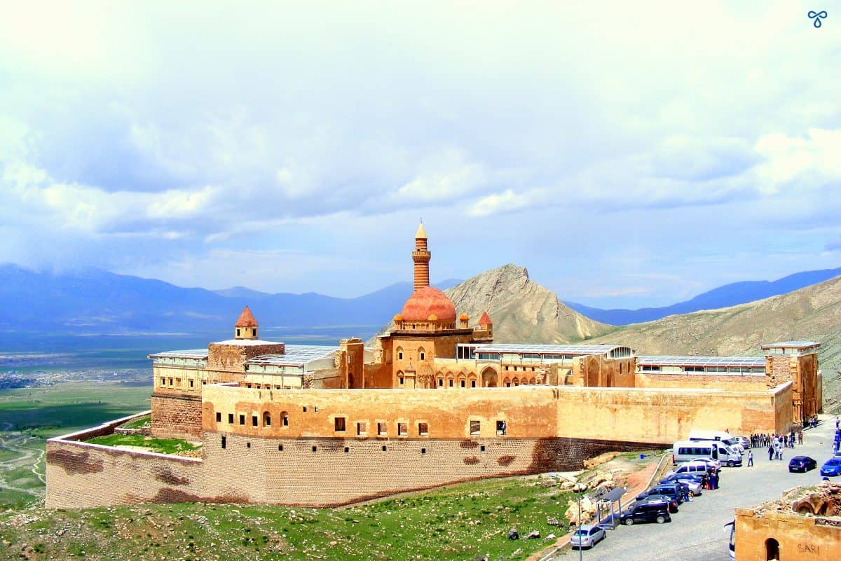 The complex of Ishak Pasha Palace from high. The stone complex is walled and the dome and minaret of the mosque can be seen. A car park is at the entrance.
