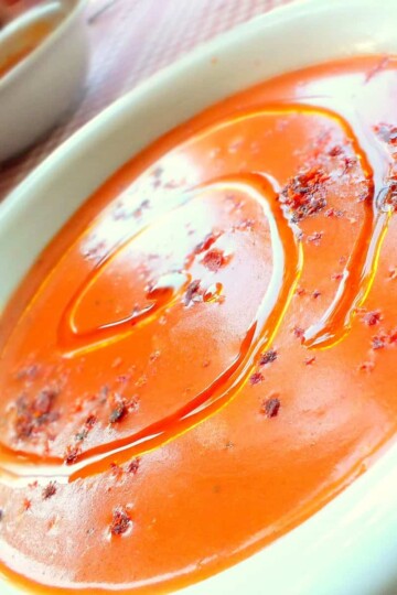 A bowl of soup on a tilted angle photo. The soup is a thick, smooth rust colour and is topped with olive oil and chilli flakes.