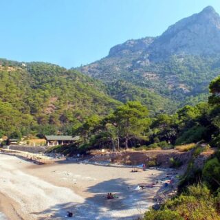 White sand and shingle of Kabak Beach backed by forested hillside and a pointed cliff. The waves are crashing on the beach.