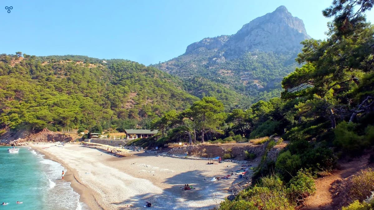White sand and shingle of Kabak Beach backed by forested hillside and a pointed cliff. The waves are crashing on the beach.