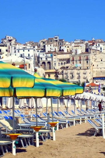 A uniform row of sun loungers with green and yellow striped parasols. A hillside town rises up the hill in the background.