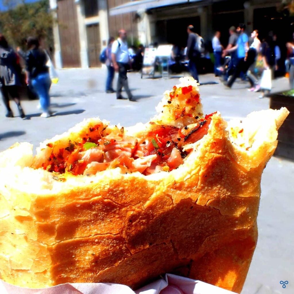 A soft crusty cob filled with chopped kokoreç. People walk by in the background.