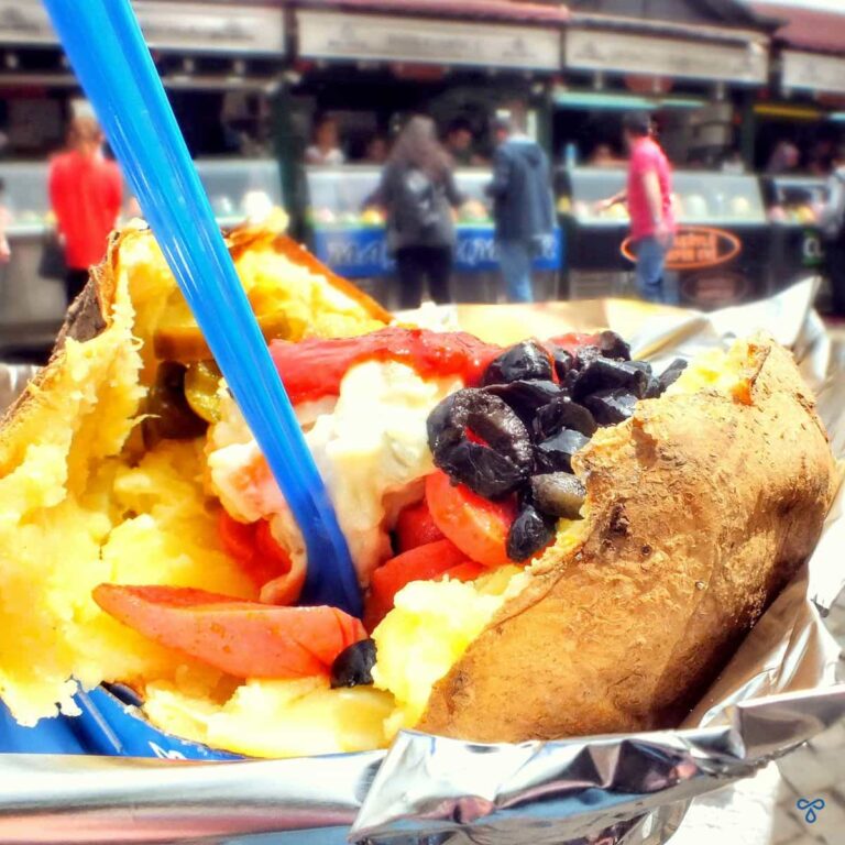 A baked potato in a foil case, topped with black olives, cocktail sausages and Russian salad