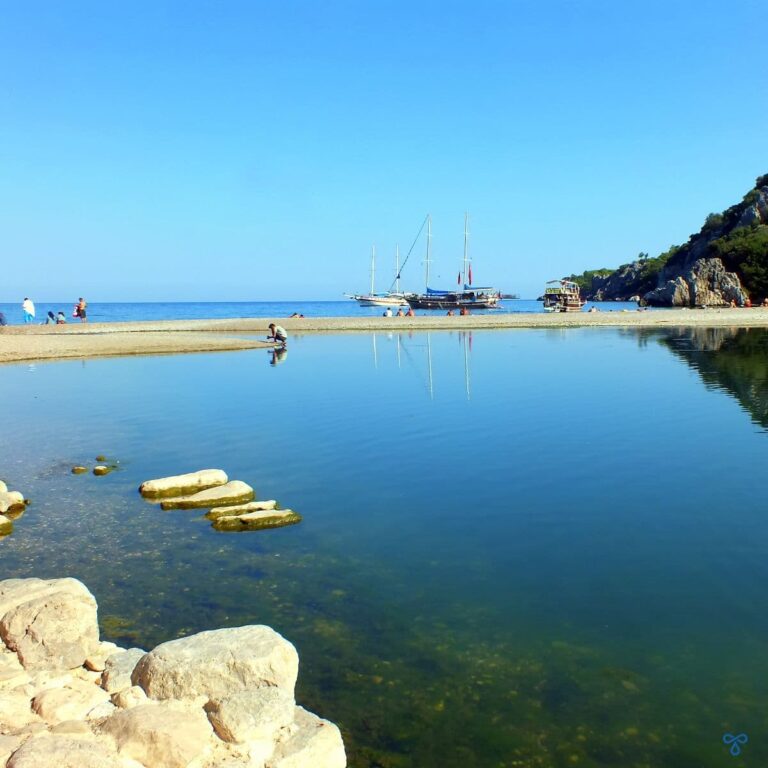 A calm lagoon with a stretch of beach in the distance and two gülets anchored along the shoreline.