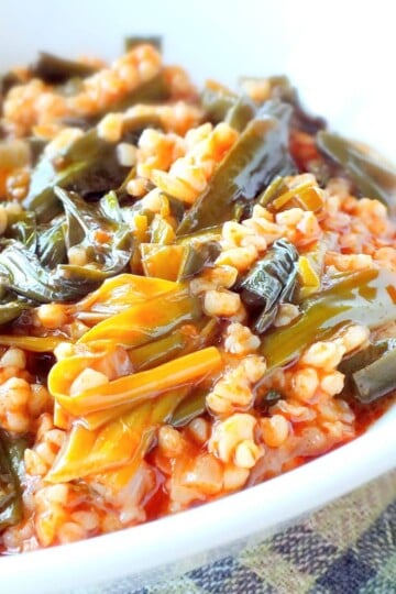 A white bowl with cooked bulgur wheat in an oil-based tomato sauce. A green leafy vegetable - çiriiş - is also in the mixture.