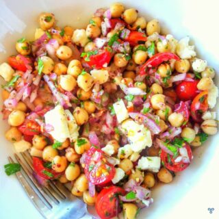A chickpea salad bowl viewed from above. Red onion slices, lumps of white cheese and tomato are in the salad.