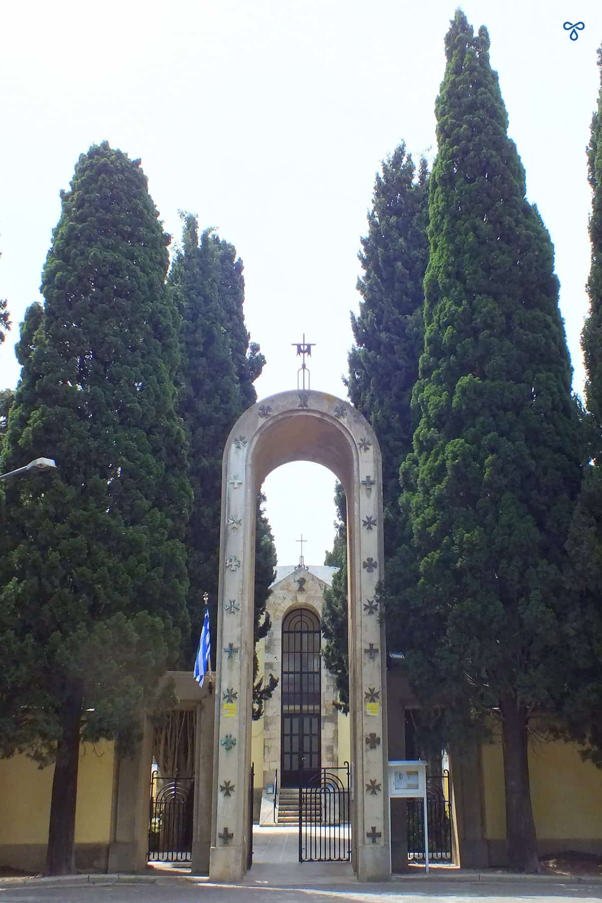 A tall arch with a cross at the top marking the entrance to Agnus Dei catholic church in Kos Town. Tall fir trees are either side of the arch.