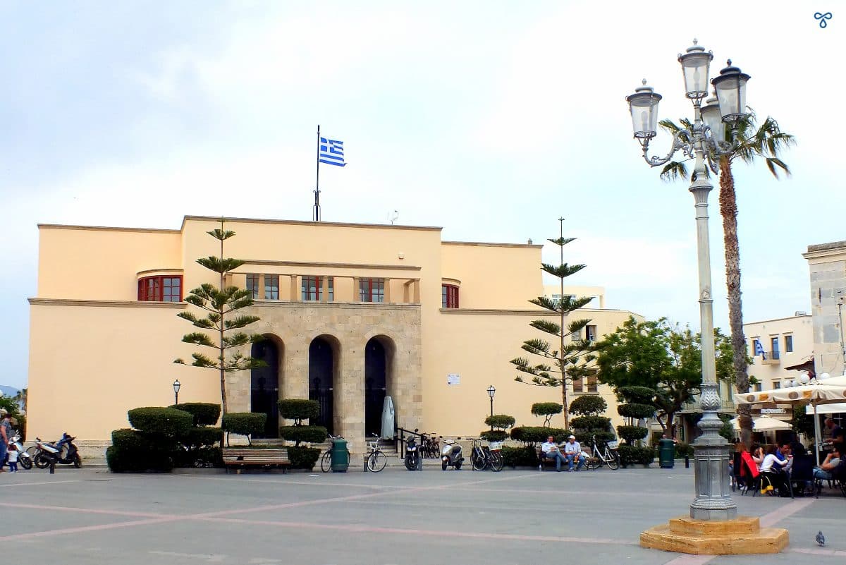 A wide art deco building with three narrow arches for the entrance. An ornate lamp is in the plaza in front of it and a Greek flag flies on top of the building. 