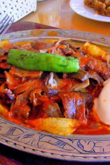 Iskender kebab - thin slices of meat in a tomato sauce. A sliced of green pepper is on top and a dollop of yoghurt on the side.
