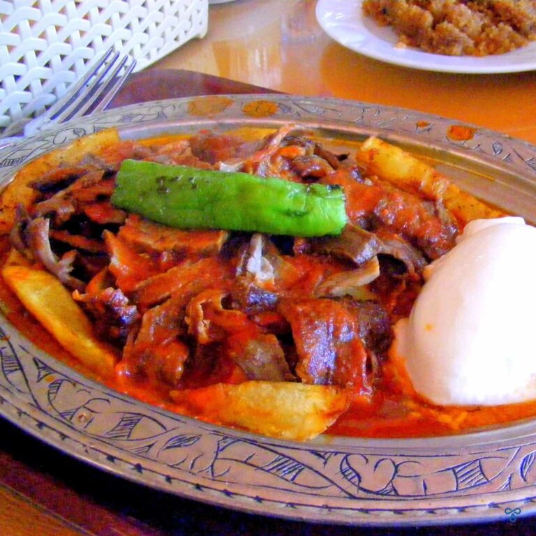 Iskender kebab - thin slices of meat in a tomato sauce. A sliced of green pepper is on top and a dollop of yoghurt on the side.