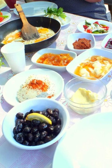 A table with various small plates making up Turkish breakfast. Fried eggs, black olives and jams are in the frame.