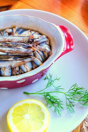 An oval shaped bowl with cooked anchovy fillets topping a dish. A slice of lemon is in the foreground.