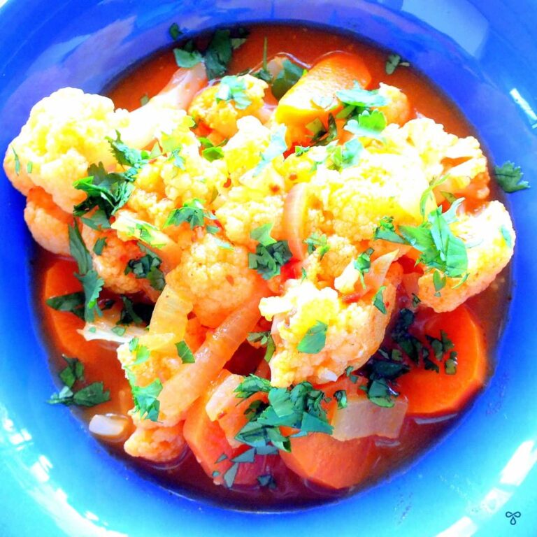 Cauliflower florets and sliced carrot in tomato gravy, stopped with fresh green chopped herbs. The meal is in a blue bowl and the photo is taken from above.