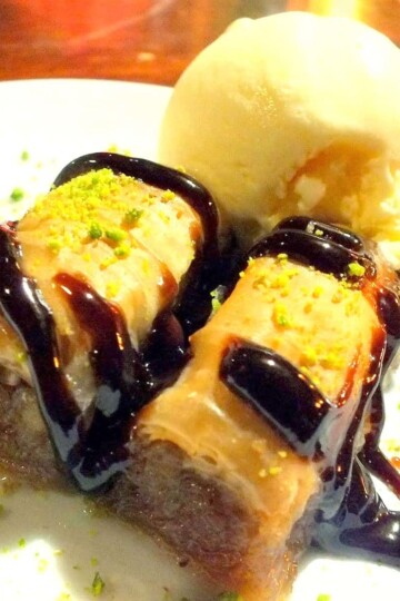 Two pieces of baklava Turkish dessert topped with crushed pistachio and chocolate sauce and served with a ball of vanilla ice cream.