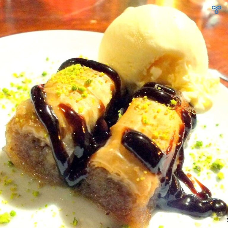 Two pieces of baklava Turkish dessert topped with crushed pistachio and chocolate sauce and served with a ball of vanilla ice cream.
