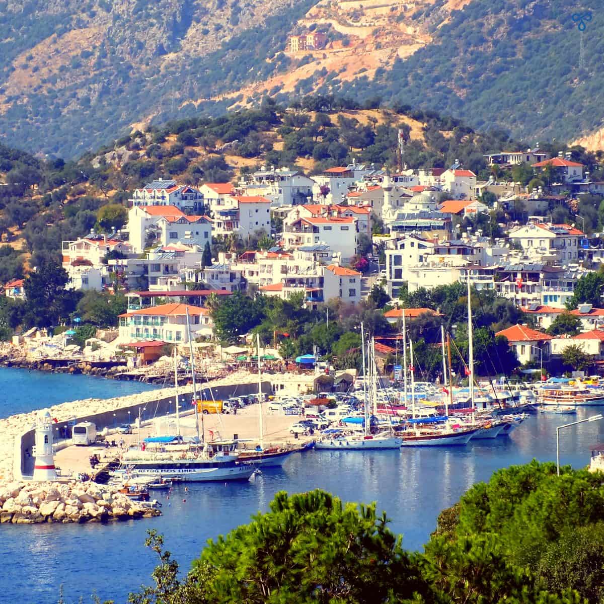 A view of Kaş harbour from a high road. Gülets and a lighthouse line the break. Whitewashed buildings climb up the hillside in the background.
