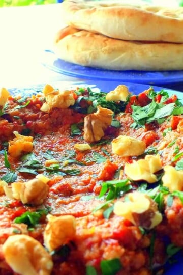 A close up of a circular blue plate with a red pepper paste, topped with broken walnuts and torn parsley leaves. Two flatbreads are in the background.