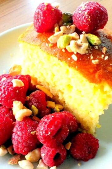 A slice of revani sponge cake topped with raspberries and broken pistachio nuts. Raspberries are also on the side.