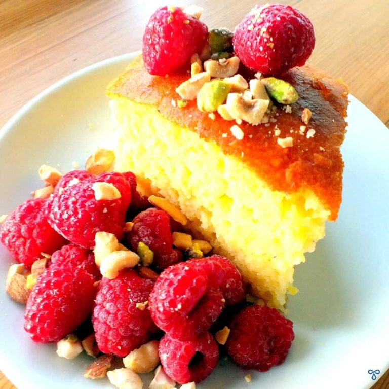 A slice of revani sponge cake topped with raspberries and broken pistachio nuts. Raspberries are also on the side.