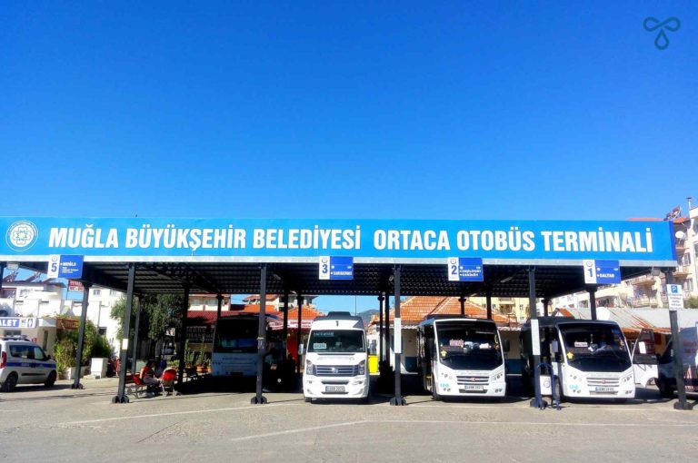 Three white minibuses under a canopy in a bus station. The canopy tells travellers they are at Ortaca Otobüs Terminali