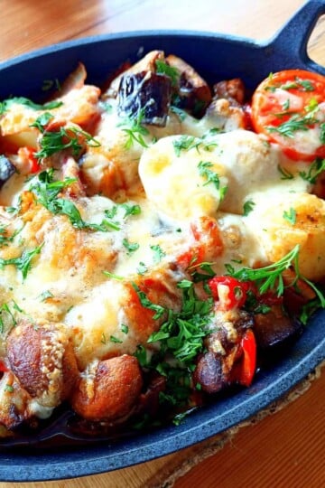 A cast iron oval bowl with cooked vegetable casserole inside. Potatoes, tomatoes and mushrooms are topped with melted cheese and chopped fresh herbs.