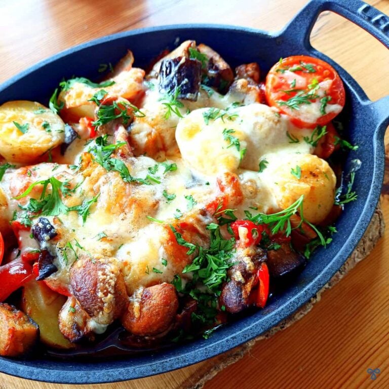 A cast iron oval bowl with cooked vegetable casserole inside. Potatoes, tomatoes and mushrooms are topped with melted cheese and chopped fresh herbs.