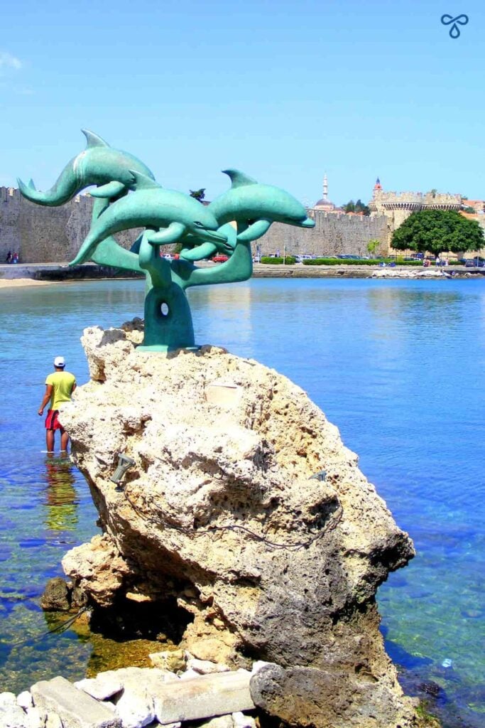 A rock stands in the sea at Rhodes harbour. It is topped by a sculpture of three green dolphins. The castle walls are in the background.