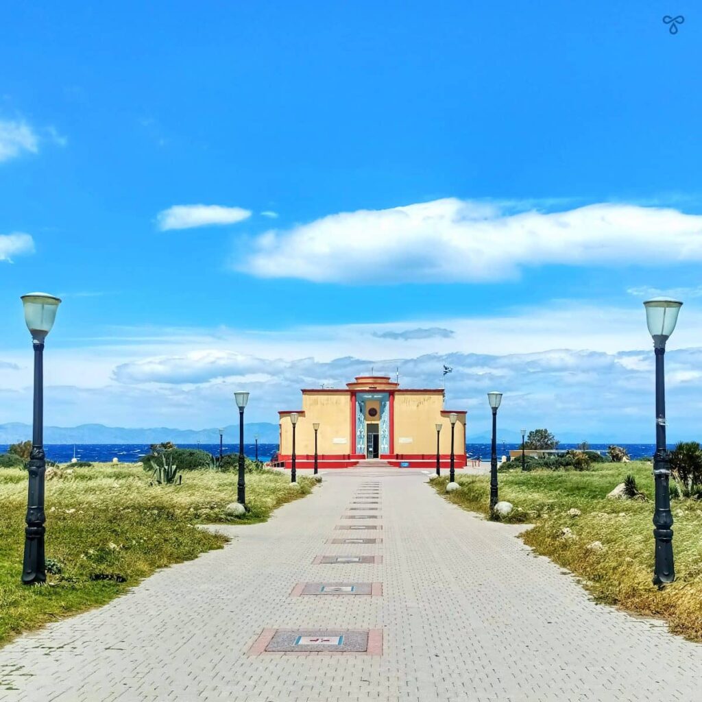 A straight path lined with streetlights, leading to the art deco Rhodes Aquarium. The sea is in the background.