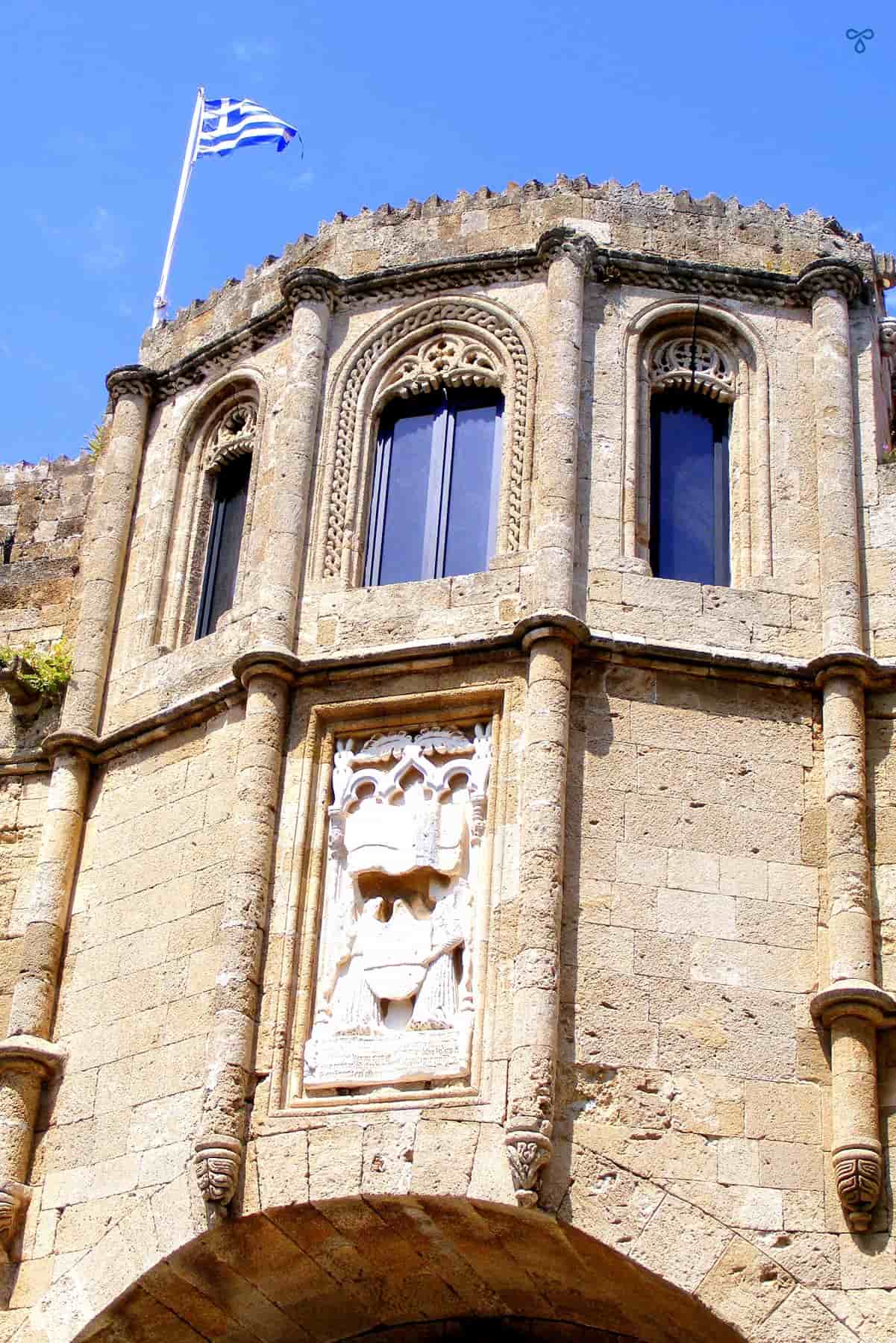 The ornate stone frontage of Rhodes Archaeological Museum. Three windows are around the top and a Greek flag flies on the roof.
