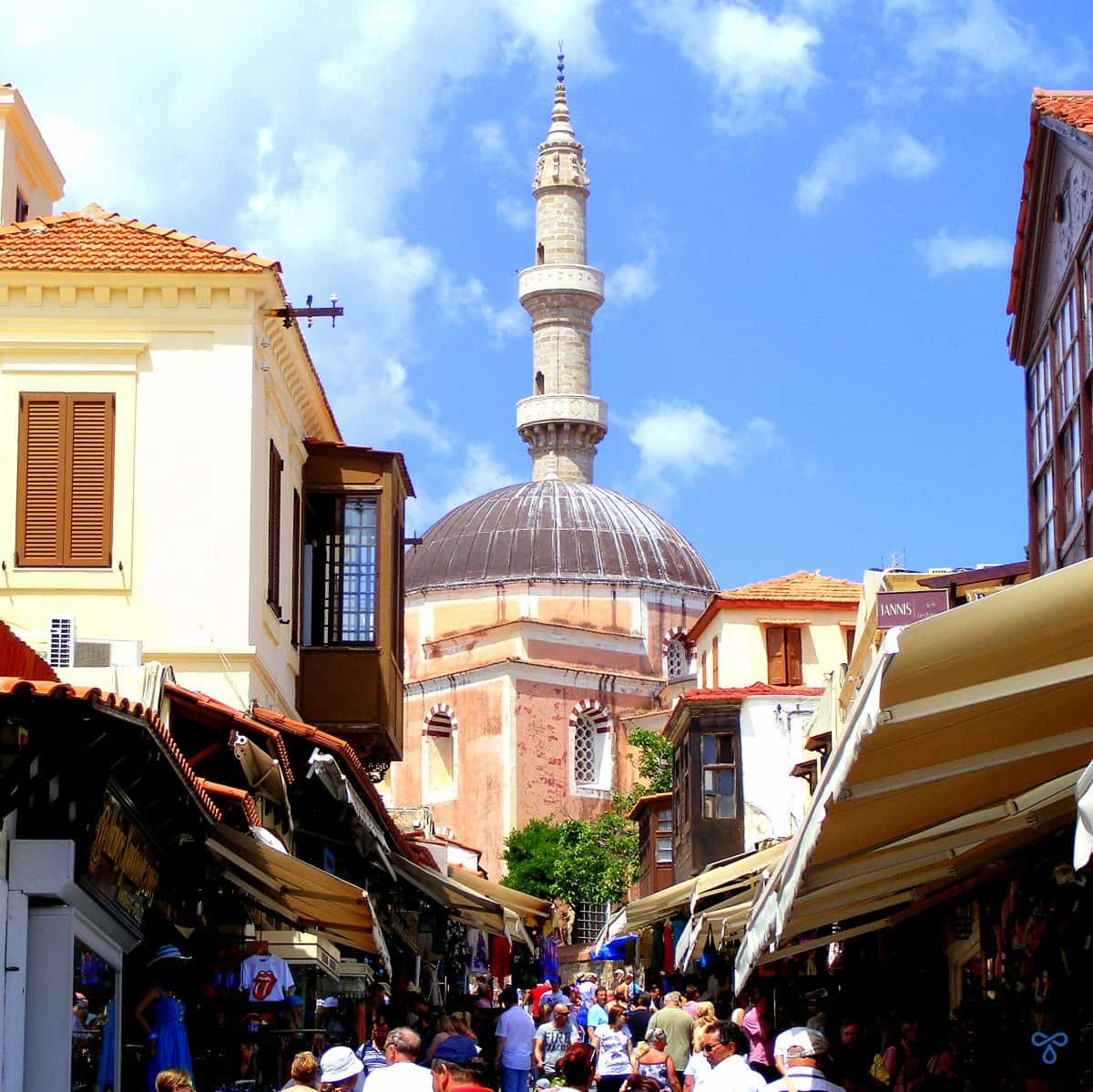 A narrow uphill, crowded, pedestrianised shopping street with the pink walls and minaret of the Mosque of Suleiman in Rhodes Old Town at the top of the hill.