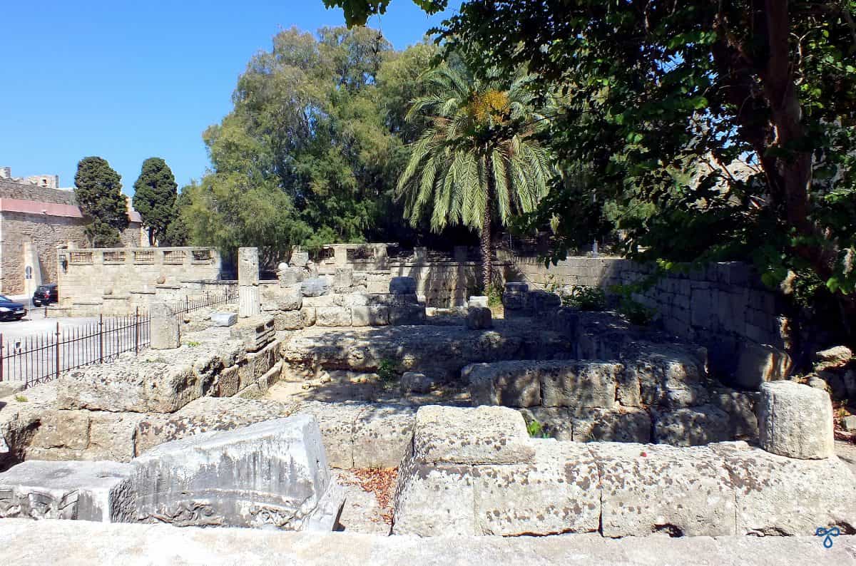 The stone ruins of the Temple of Aphrodite in Rhodes Old Town. Partial columns and a palm tree stand amongst the large stones.