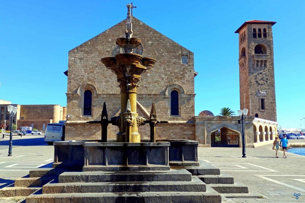 A stone church with a separate clock tower to the right and a fountain in front of in Rhodes Town.