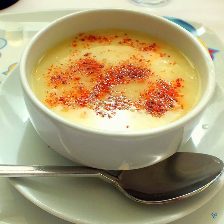 A bowl of yellow soup topped with red chilli flakes. The soup bowl is on a plate with a spoon.