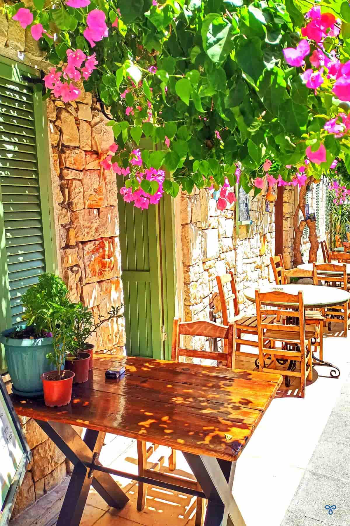 A portrait shot of a stone building shaded by bougainvillea. Wooden chairs and tables are outside.