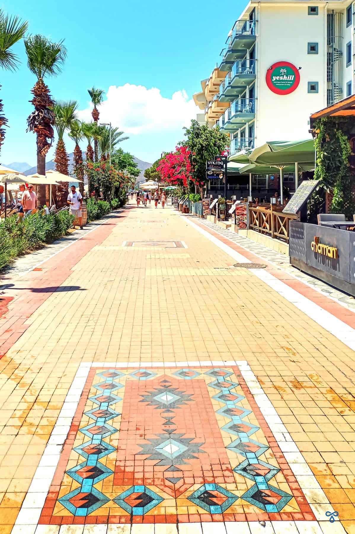 A beach promenade with patterned paving. Bars and restaurants line the path.